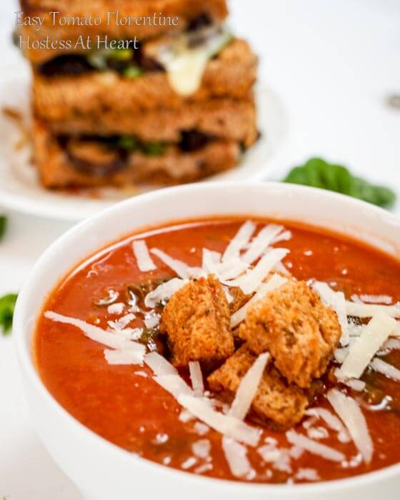 A white bowl of tomato Florentine soup garnished with shredded cheese and croutons. A grilled cheese sits in the background.