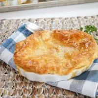 A pot pie topped with a puff pastry crust sitting in a white dish over a blue-checked napkin. More pies sit on a baking sheet in the background.