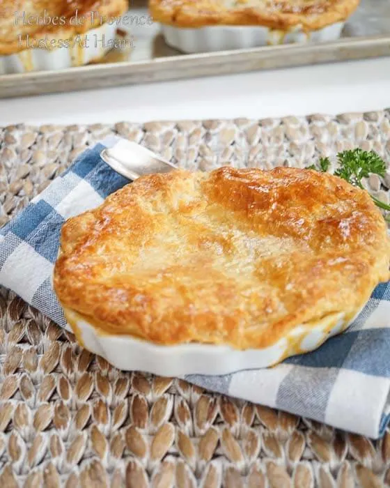 A pot pie topped with a puff pastry crust sitting in a white dish over a blue-checked napkin. More pies sit on a baking sheet in the background.