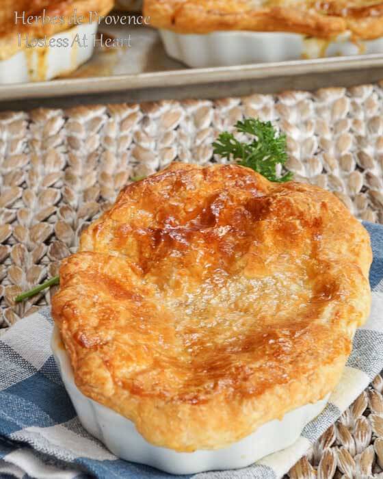 A pot pie topped with a puff pastry crust sitting in a white dish over a blue-checked napkin. More pies sit on a baking sheet in the background.