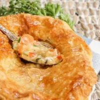 A pot pie topped with a puff pastry crust sitting in a white dish over a blue-checked napkin. A spoon of the filling hovers over the pie.