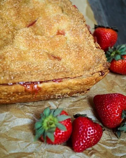 Square slab Strawberry pie sitting on a piece of parchment surrounded by fresh strawberries. 