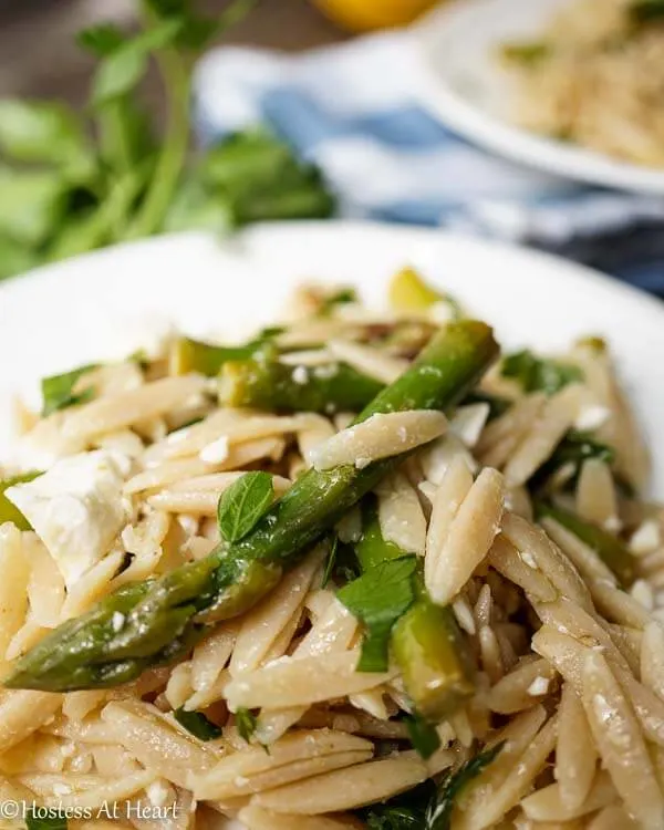 A white plate filled with orzo and asparagus and garnished with feta. A second plate sits in the background over a blue-checked napkin.A plate of food, with Salad and Orzo