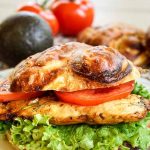 A grilled chicken sandwich on a pretzel bun layered with lettuce and tomato. Fresh tomatoes and an avocado sits in the background.
