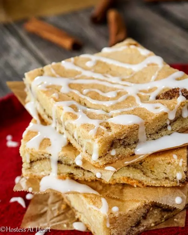 A stack of Snickerdoodle Bars with glaze running down the sides over a red napkin. Cinnamon sticks lay in the background.