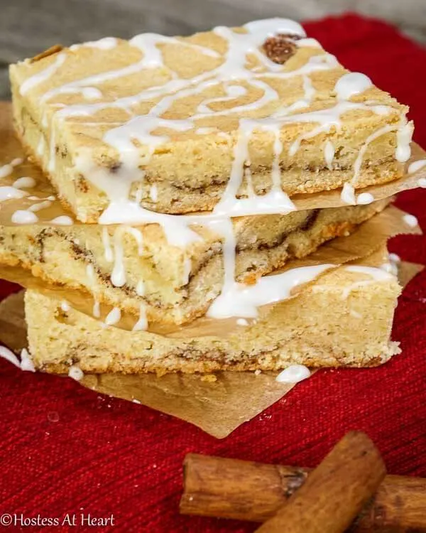 A stack of Snickerdoodle Bars with glaze running down the sides over a red napkin. Cinnamon sticks lay in the background.