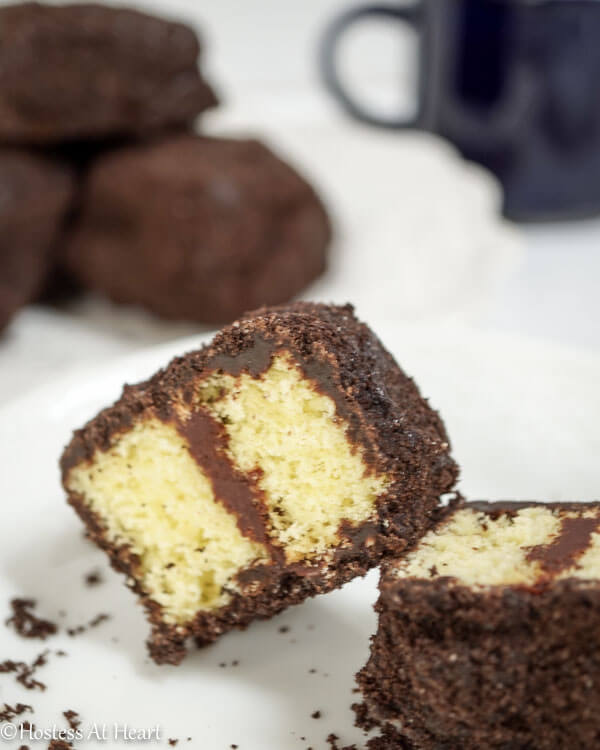 A plate with a chocolate lamington cut in half showing yellow cake layered with chocolate frosting. A plate of lamingtons sit in the background.