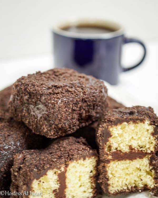 A plate of chocolate lamingtons in front of a cup of coffee. Two of the lamingtons are cut in half showing yellow cake layered with chocolate frosting.