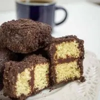 A plate of chocolate lamingtons in front of a cup of coffee. Two of the lamingtons are cut in half showing yellow cake layered with chocolate frosting.