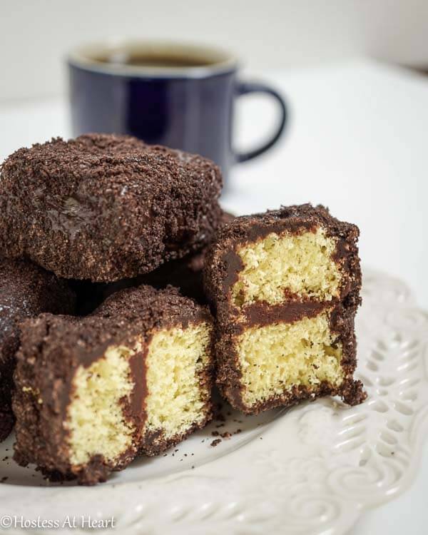 A plate of chocolate lamingtons in front of a cup of coffee. Two of the lamingtons are cut in half showing yellow cake layered with chocolate frosting.