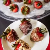 Top view of 4 chocolate coated strawberries on a white plate in from of platter of more berries