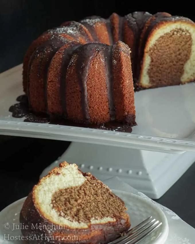 Slice of a two-toned cake on a plate in front of the whole cake sitting on a cake stand in the background.