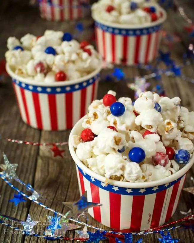 Angled view of a bowl of candied popcorn dotted with red and blue chocolate candy. A string of stars weaves around the bowl with two bowls in the background. 