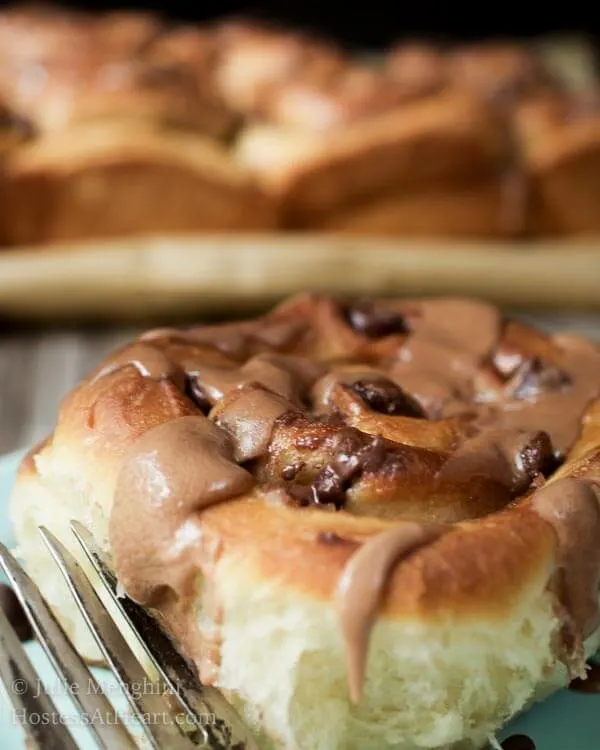 A sweet roll topped with chunks of chocolate and a malt-flavored icing on a light blue plate over a wooden board. A fork sits next to the roll. The pan of rolls sits in the background.