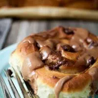 A sweet roll topped with chunks of chocolate and a malt-flavored icing on a light blue plate over a wooden board. The pan of rolls sits in the background.