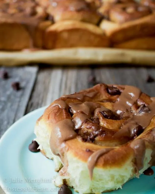 A sweet roll topped with chunks of chocolate and a malt-flavored icing on a light blue plate over a wooden board sprinkled with additional chocolate pieces. The pan of rolls sits in the background.