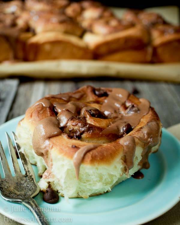 A sweet roll topped with chunks of chocolate and a malt-flavored icing on a light blue plate next to a fork over a wooden board. The pan of rolls sits in the background.