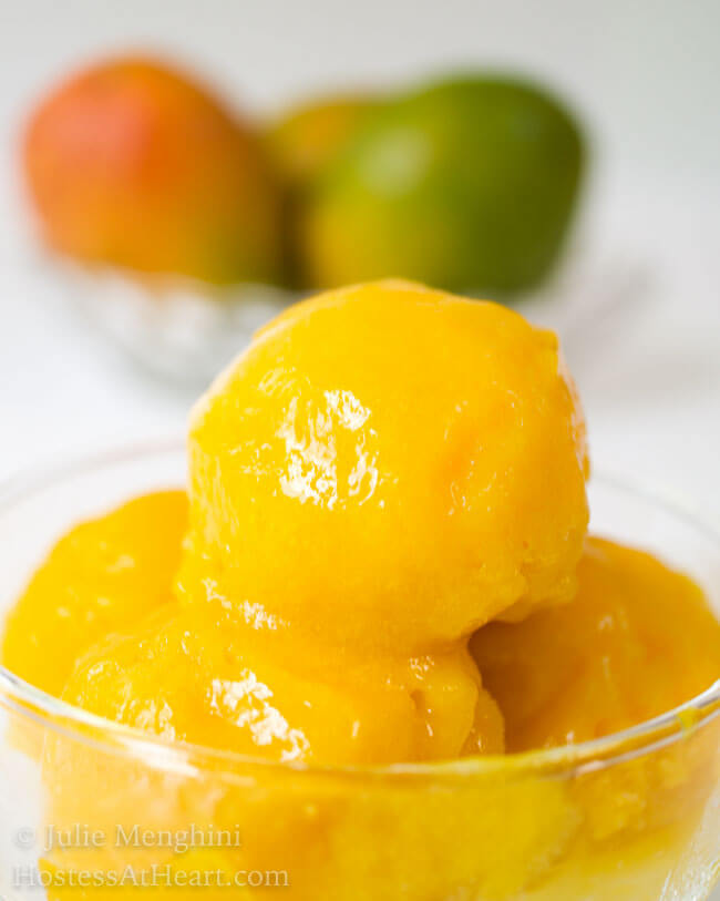 Scoops of mango sorbet in a glass bowl sitting in front of fresh mangos.