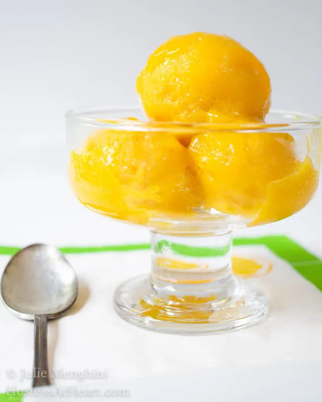 Scoops of mango sorbet in a glass bowl sitting in front of fresh mangos.