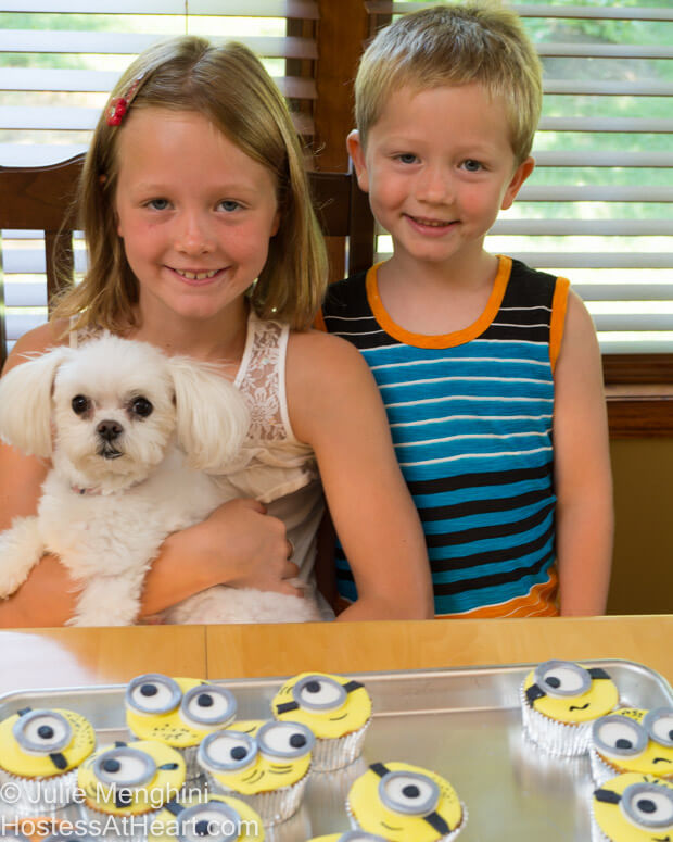 Two children and a dog sitting in front of minion cupcakes.