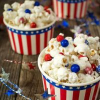 Angled view of a bowl of candied popcorn dotted with red and blue chocolate candy. A string of stars weaves around the bowl with two bowls in the background.