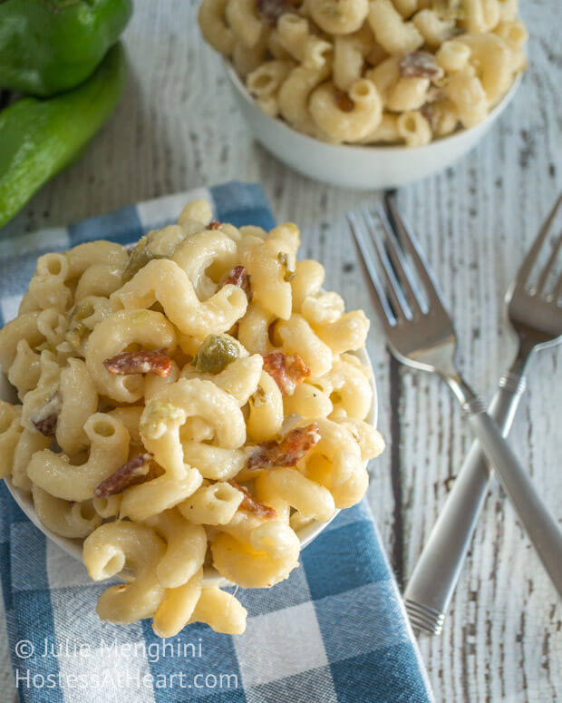 Top-down view of a bowl of hatch chili & bacon macaroni and cheese over a blue checked napkin. Forks and another bowl sit off to the side.