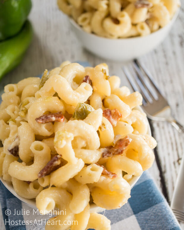 Top-down view of a bowl of hatch chili & bacon macaroni and cheese over a blue checked napkin. Forks and another bowl sit off to the side.