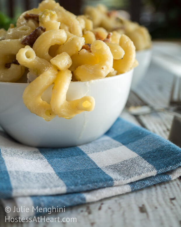 A close-up side view of macaroni and cheese with hatch chilies and bacon in a white bowl sitting on a blue checked napkin.