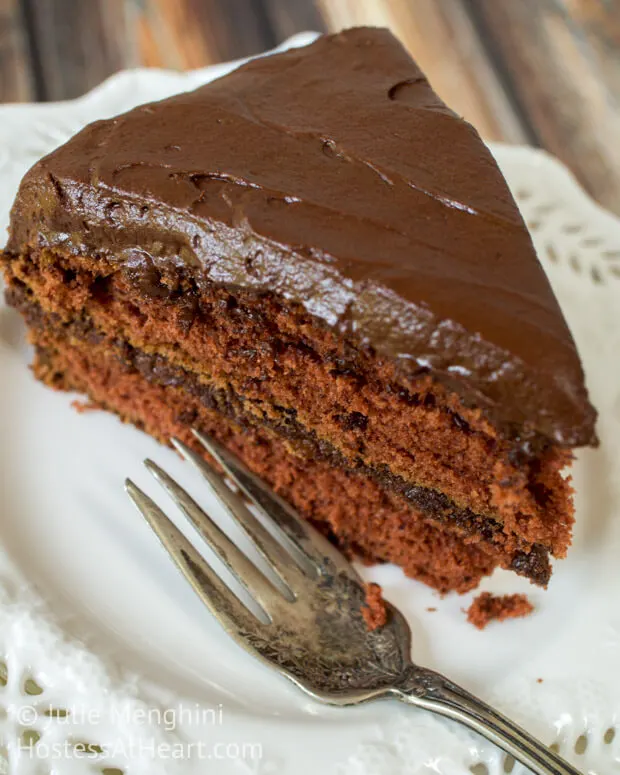 Top-down view of a slice of chocolate layered cake sitting on a white plate. A fork sits on the plate.