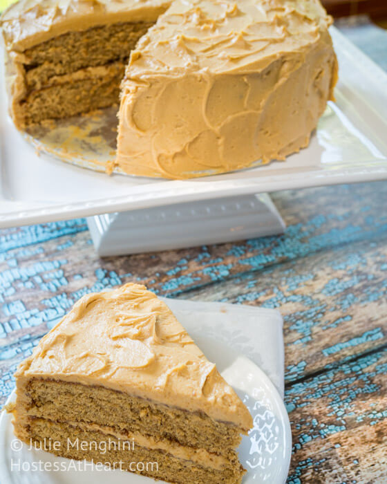 A piece of layered Spice Cake with Peanut Butter frosting between the layers and over the top on a white plate in the forefront. The round cake on a cake pedestal is in the background.