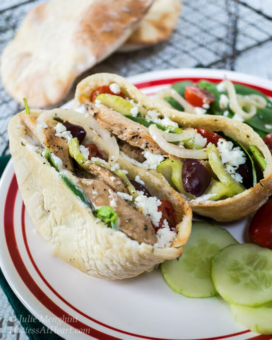 Two open pita halves filled with sliced chicken, onion kalamata olives, cucumbers, and Feta cheese sits on a white plate. Cooked pitas sit on a cooling rack in the back.