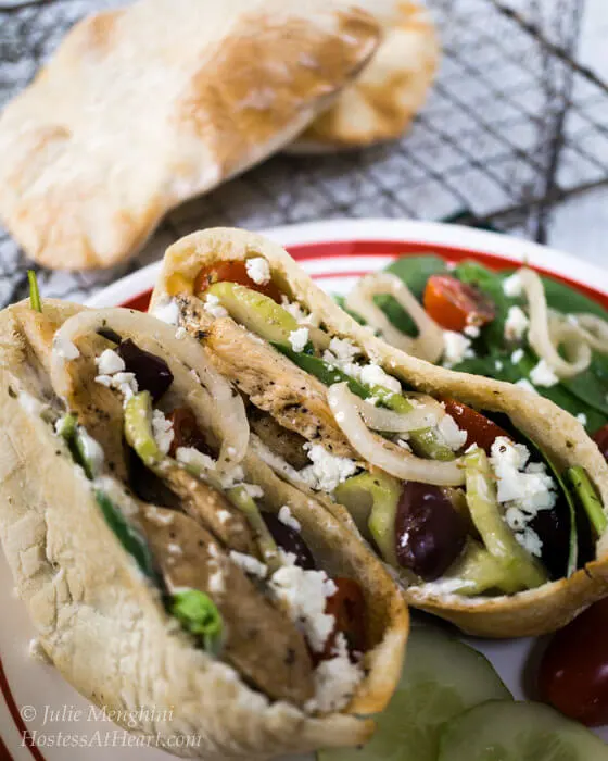 A pita sliced in half and stuffed with Chicken Gyro ingredients including chicken onions, kalamata olives, cucumbers, and feta cheese. Cooked pita shells sit in the background on a cooling rack.