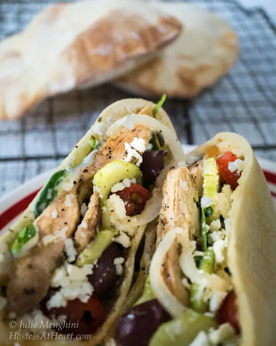 A pita sliced in half and stuffed with Chicken Gyro ingredients including chicken onions, kalamata olives, cucumbers, and feta cheese. Cooked pita shells sit in the background on a cooling rack.