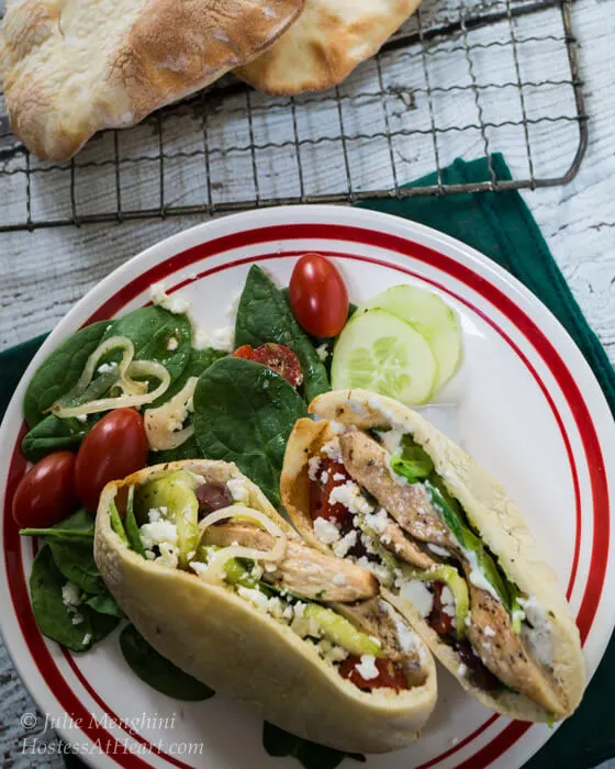 A pita sliced in half and stuffed with Chicken Gyro ingredients including chicken onions, kalamata olives, cucumbers, and feta cheese sitting on a white plate next to a spinach salad. Cooked pita shells sit in the background on a cooling rack.