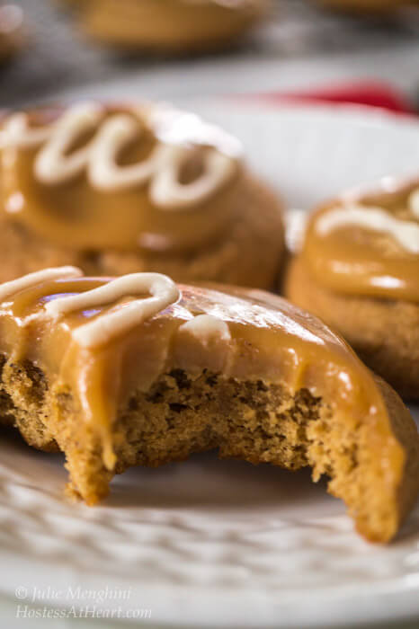 A close up 3 cookies topped with caramel and a white swirl on a white plate. The front cookie has a large bite out of it showing the soft center and thick caramel topping.