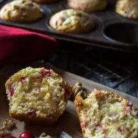 A cranberry-Orange muffin that's been cut in half showing it's the soft center. A muffin tin sits in the back filled with more muffins.
