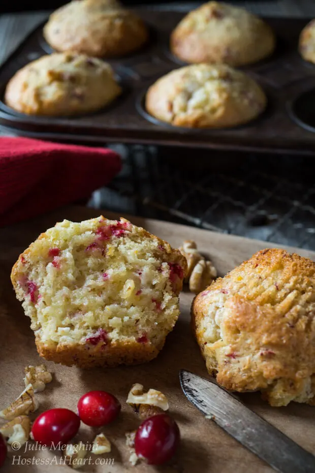 A cranberry-Orange muffin that\'s been cut in half showing it\'s the soft center. A muffin tin sits in the back filled with more muffins.