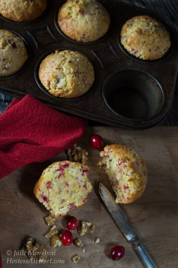 A cranberry-Orange muffin that\'s been cut in half showing it\'s the soft center. A muffin tin sits in the back filled with more muffins.