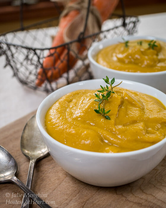 Two white bowls of creamed vegetable soup with a wire basket containing carrots in the background.