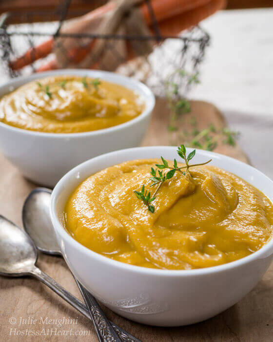 Two white bowls of creamed vegetable soup with a wire basket containing carrots in the background.