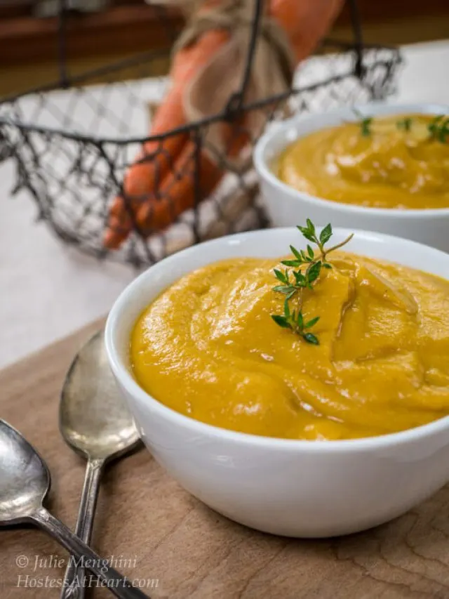 Two white bowls of creamed vegetable soup with a wire basket containing carrots in the background.