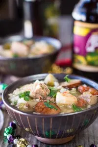 A bowl Gumbo loaded with Shrimp, Andouille Sausage, and Duck. A second bowl and a beer sit behind it.