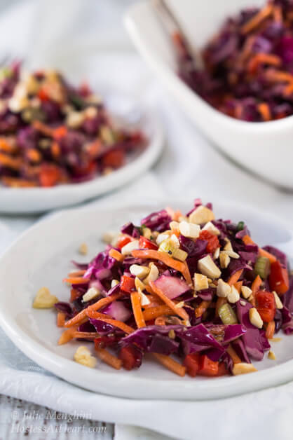 A top-angled Asian Cabbage Salad filled with chopped red cabbage, cashews, carrots, and red peppers in a sweet ginger sesame vinaigrette sitting on a white dish. A bowl and plate filled with the salad sit in the background.