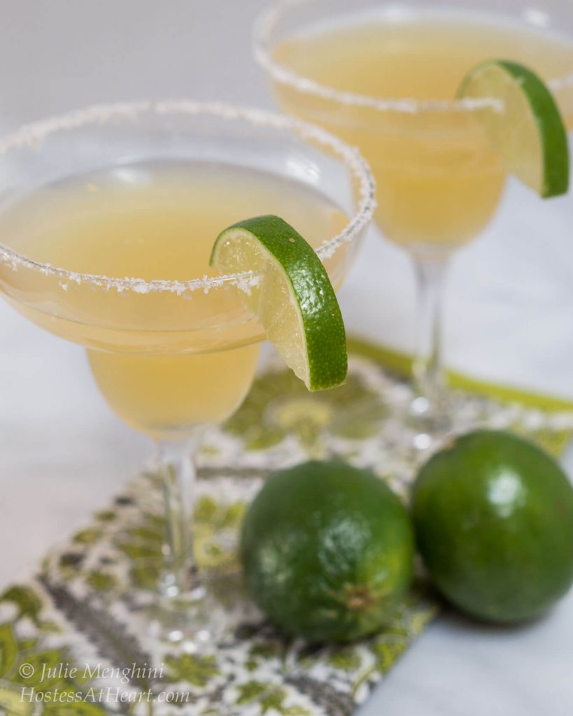 Two margarita glasses rimmed with salt and filled with margaritas. Two fresh limes sit in the front.