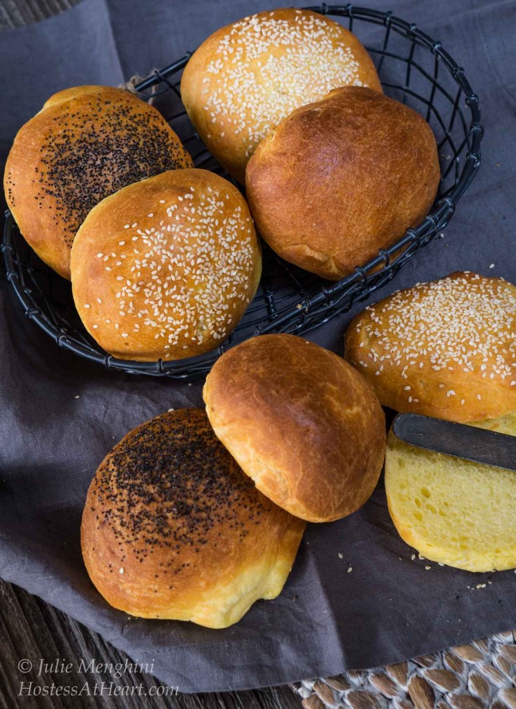 Top-down view of brioche buns sitting in a wire basket and on a piece of slate. Some buns are plain. Others are topped with poppyseed or sesame seeds.