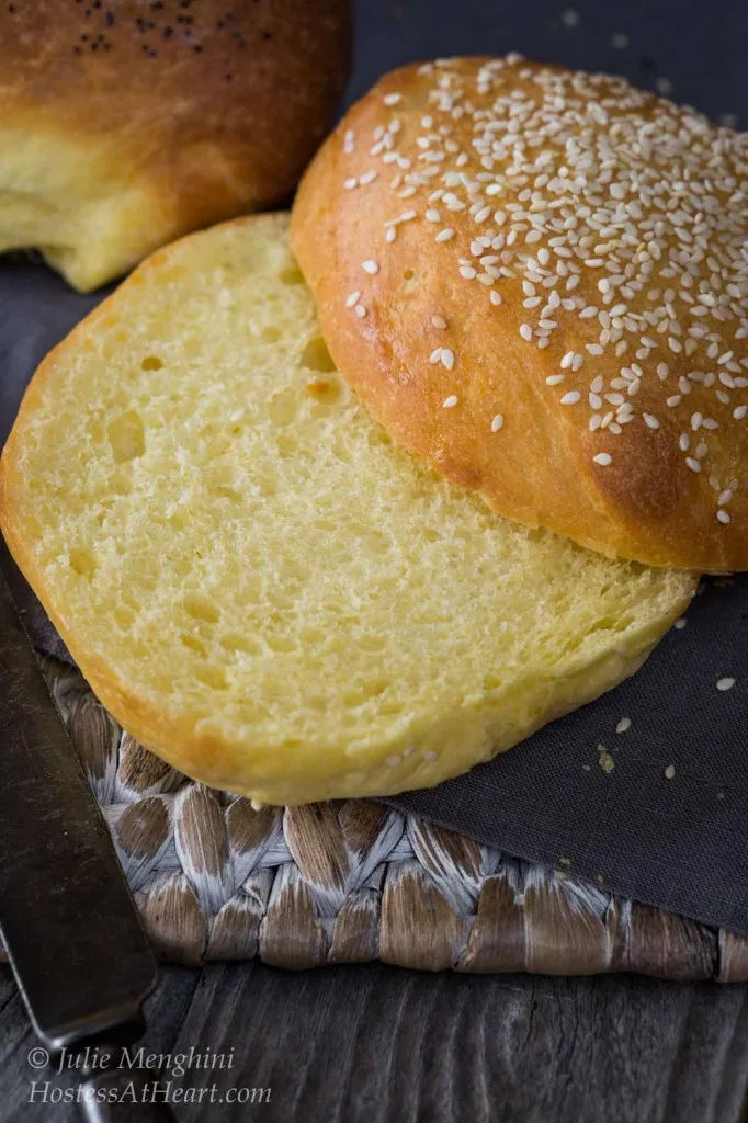 A closeup view of a sliced brioche bun topped with sesame seeds. More buns sit in the background.