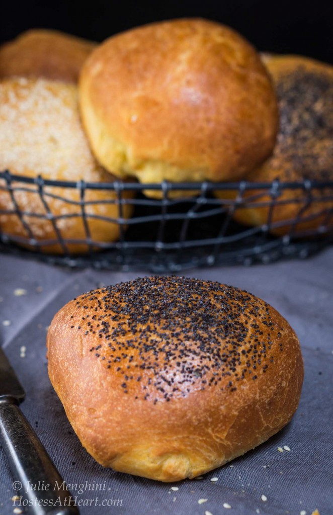 A Broche Bun topped with poppy seeds sits on a blue napkin in front of a basket of Brioche rolls.