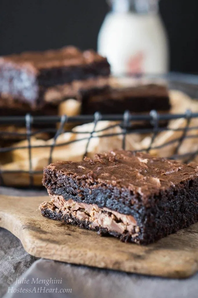 Slice of Dark chocolate brownie filled with a light chocolate filling sitting on a wooden paddle. A basket filled with cut brownies sits in the background.