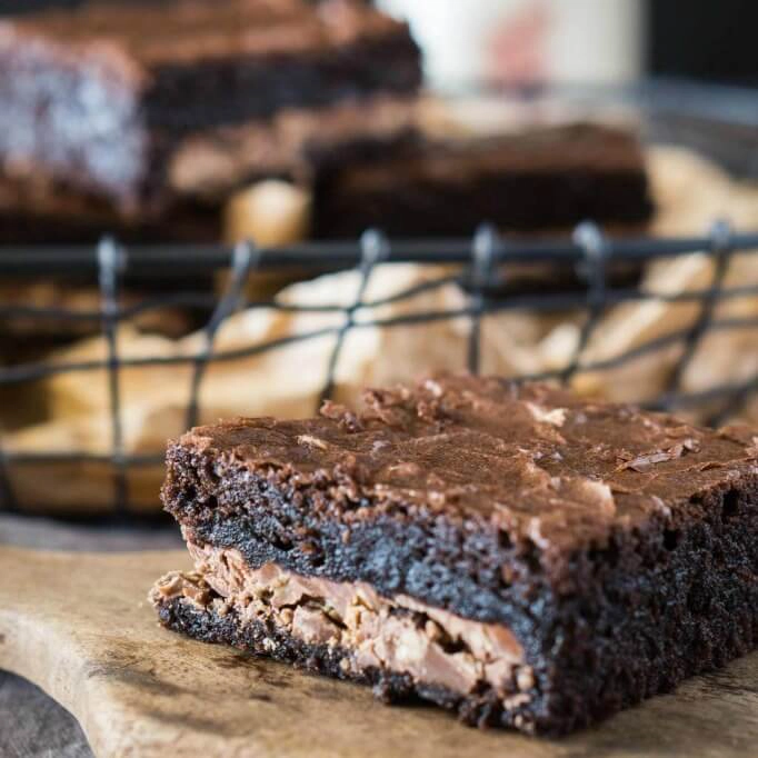 A sideview of a chocolate toffee bar sitting on a wooden paddle with a wire basket of bars sitting in the background.