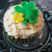  Cupcake decorated with a basketweave pattern. A green Shamrock and flower sit on the top. The cupcake sits on a metal plate.